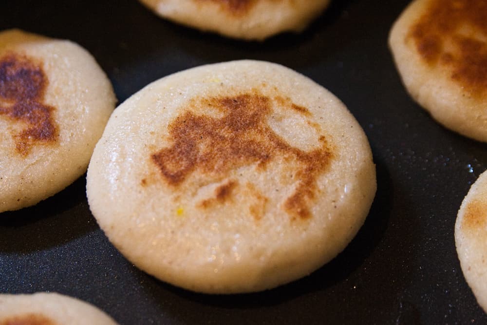 Several golden arepas cooking on a griddle, some with melted cheese